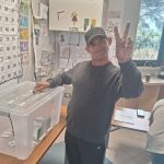 Hombre adulto en Casa de Acogida ejerciendo su voto asistido en elecciones, sonriendo y haciendo el signo de la paz junto a la urna transparente