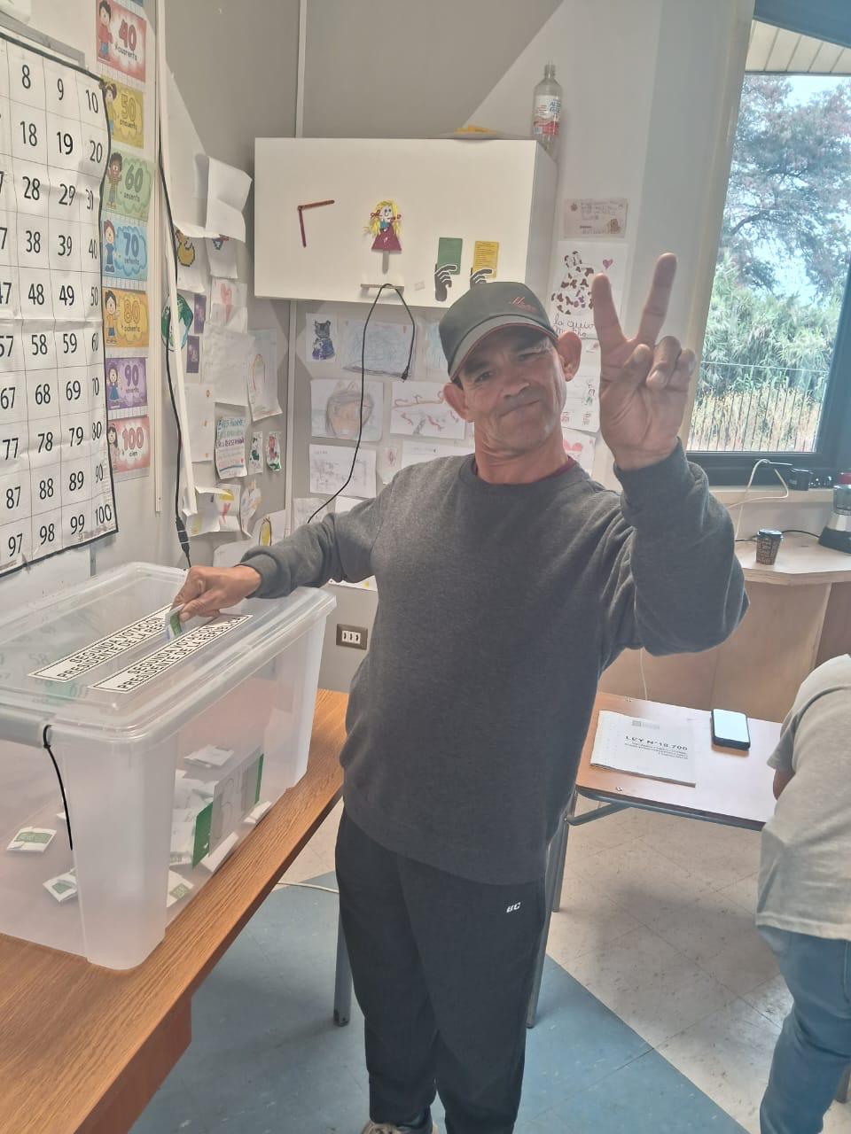 Hombre adulto en Casa de Acogida ejerciendo su voto asistido en elecciones, sonriendo y haciendo el signo de la paz junto a la urna transparente